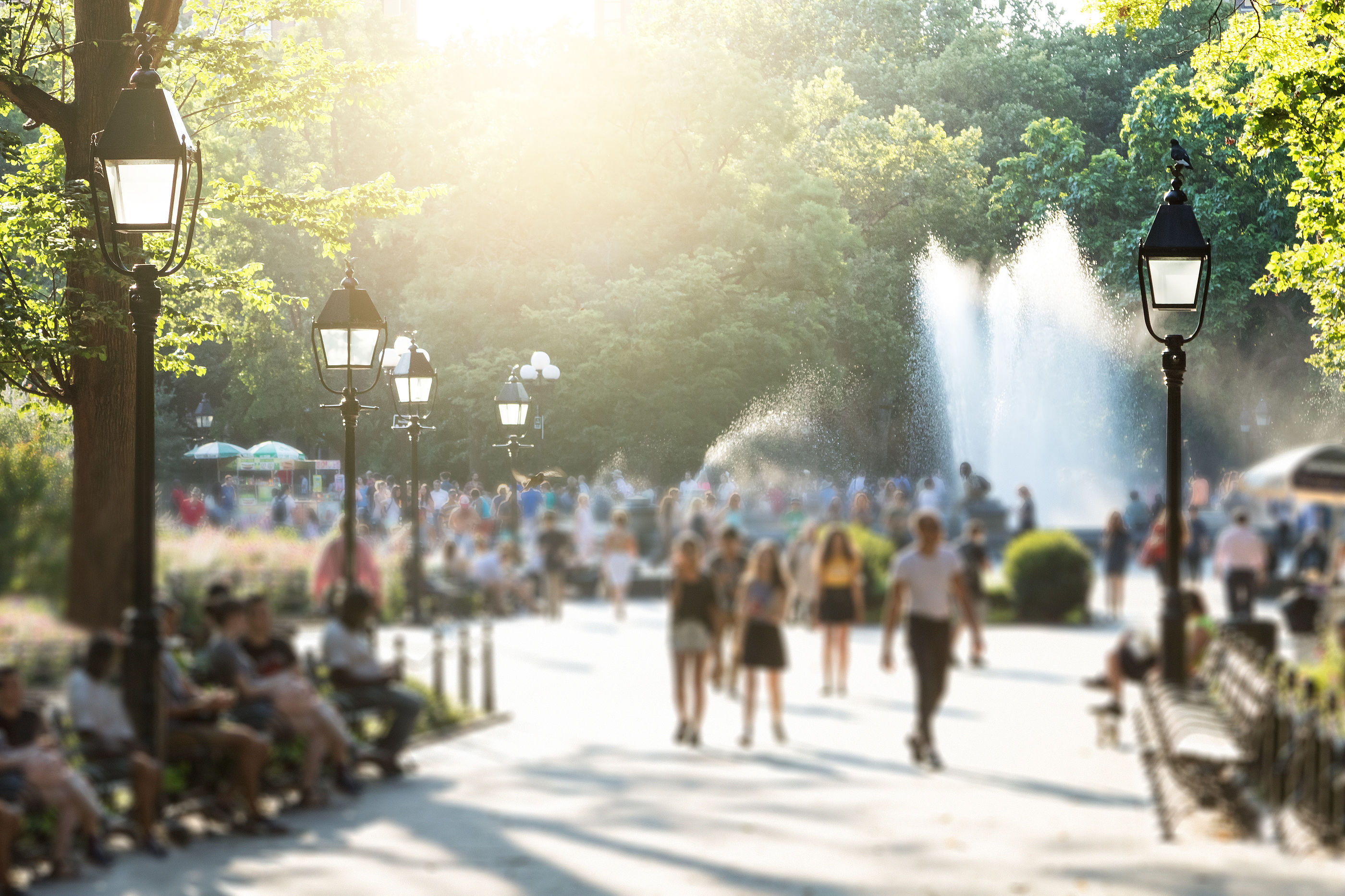 Anonymous crowds of people walking in Washington Square Park in New York City with the glow of sunlight shining in the background